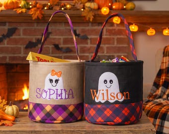 Puede incluir: Dos cubos cilíndricos para dulces de Halloween con asas. El cubo izquierdo es color canela con un fantasma con un lazo naranja y el nombre Sophia en morado. El cubo derecho es negro con un fantasma y el nombre Wilson en naranja. Bases a cuadros.