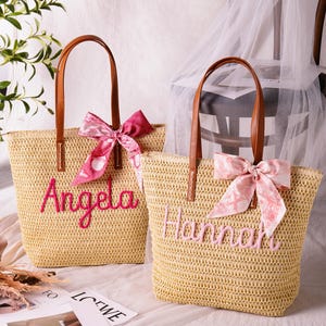 May include: Two woven straw tote bags with brown leather-look handles. One bag features the name Angela in bright pink embroidery with a dark pink patterned ribbon. The other bag displays the name Hannah in light pink embroidery with a light pink ribbon.