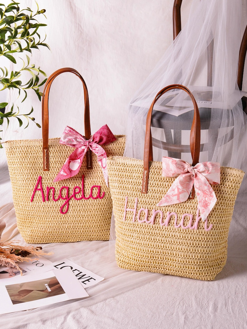 May include: Two woven straw tote bags with brown leather-look handles. One bag features the name Angela in bright pink embroidery with a dark pink patterned ribbon. The other bag displays the name Hannah in light pink embroidery with a light pink ribbon.