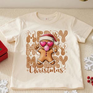 May include: Cream-colored t-shirt with a gingerbread man graphic wearing heart-shaped sunglasses and a Santa hat. The text "MERRY CHRISTMAS" is displayed in a festive font. A Christmas-themed tee.
