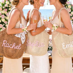 May include: Three women holding champagne flutes and personalized tote bags at a beach wedding. The bags are woven with the names "Rachel", "Sophia", and "Emily" in pink and white script. The bags have lace and floral accents.