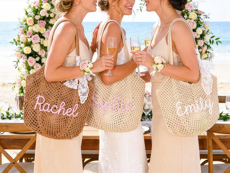 May include: Three women holding champagne flutes and personalized tote bags at a beach wedding. The bags are woven with the names "Rachel", "Sophia", and "Emily" in pink and white script. The bags have lace and floral accents.