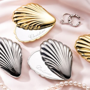 Könnte beinhalten: Eine weiße Keramiktasse mit schwarzem Henkel und dem Text "But First, Coffee" in fetter, schwarzer Schrift. Die Tasse steht vor einem verschwommenen Hintergrund, der eine gemütliche Umgebung suggeriert.