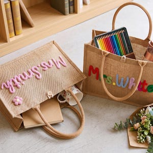 May include: Two rectangular tan jute tote bags with rope handles and button closures. One bag features the pink embroidered text Mrs. Smith and a small pink flower. The other bag displays the multicolored embroidered text Mrs. White.
