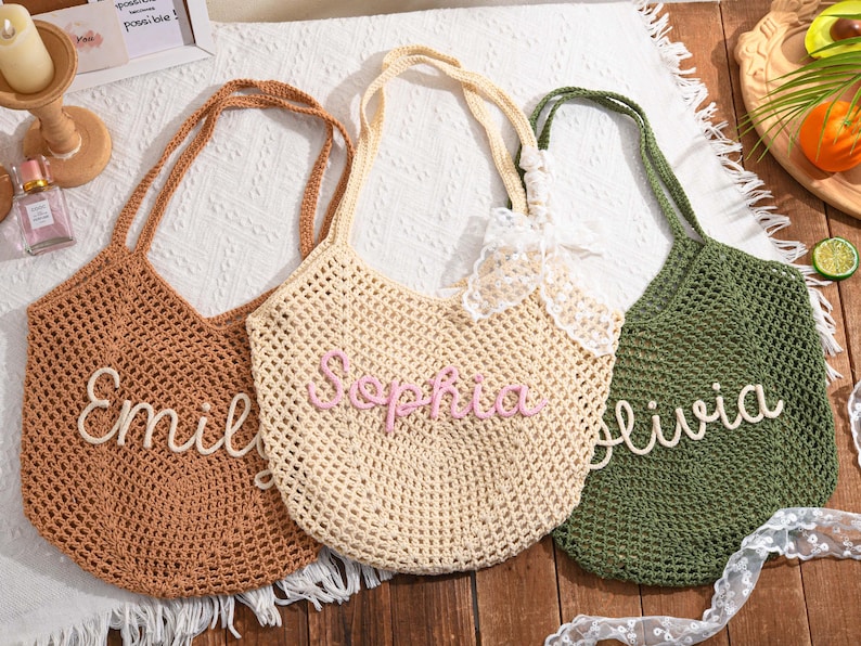 May include: Three crochet tote bags in brown, beige, and green. Each bag features a name in a contrasting color: "Emily", "Sophia", and "Olivia". The bags have long handles and a net-like weave.