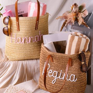 May include: Two woven straw tote bags with brown leather handles. The left bag features the name Hannah in pink embroidery, while the right bag displays the name Regina in white embroidery. Both bags contain striped towels, tumblers, and sunglasses.