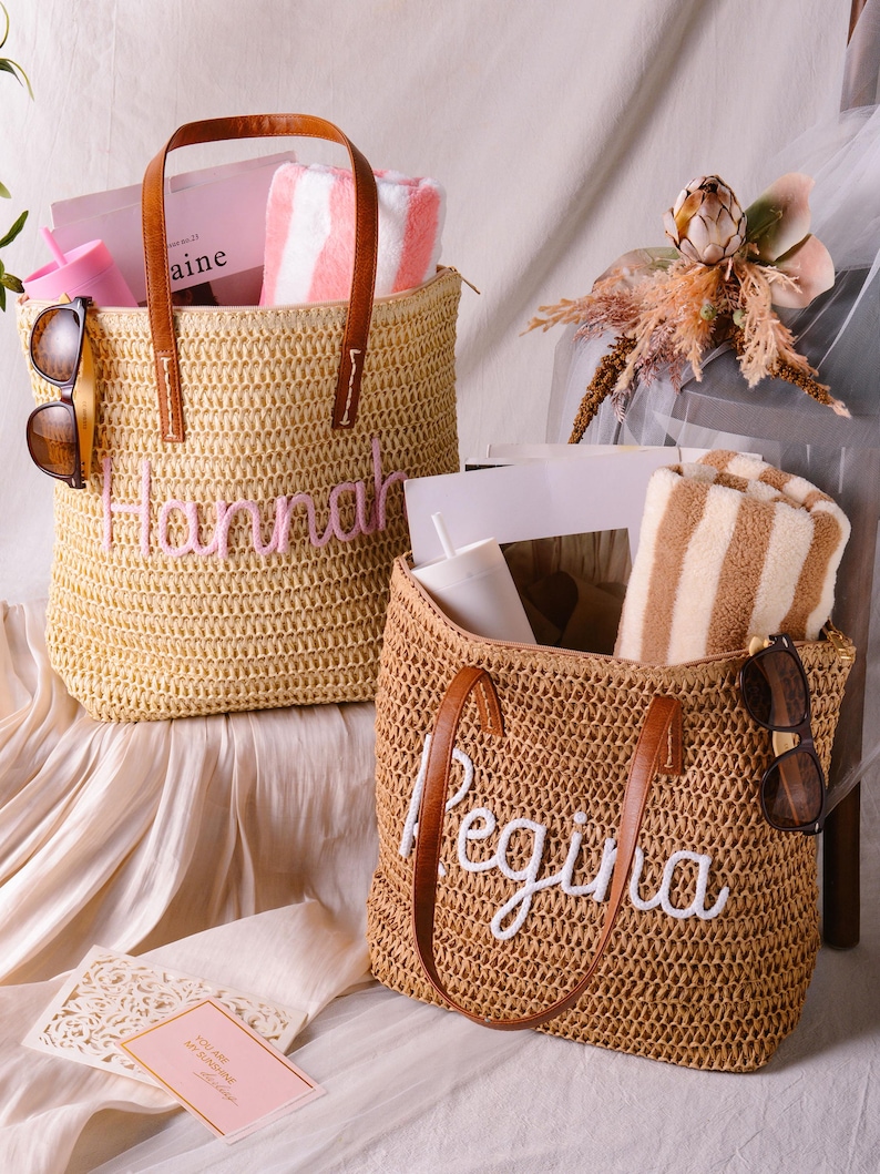May include: Two woven straw tote bags with brown leather handles. The left bag features the name Hannah in pink embroidery, while the right bag displays the name Regina in white embroidery. Both bags contain striped towels, tumblers, and sunglasses.