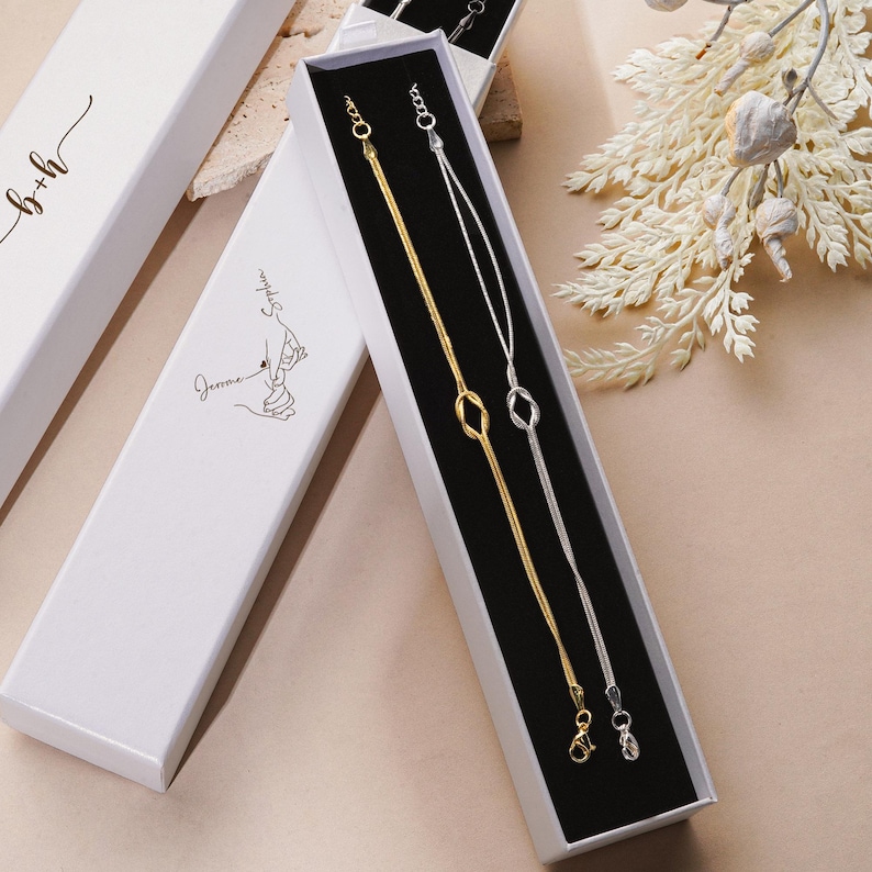 K&ouml;nnte beinhalten: Zwei feine Halsketten, eine goldene und eine silberne, pr&auml;sentiert in einer wei&szlig;en Geschenkbox mit schwarzem Innenraum. Die Halsketten haben ein Kettenmuster mit einem knotenf&ouml;rmigen Anh&auml;nger. Die Box ist ge&ouml;ffnet und zeigt den Schmuck. Weitere wei&szlig;e Boxen sind sichtbar.