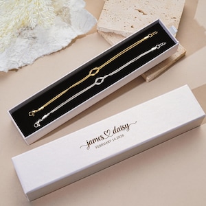 K&ouml;nnte beinhalten: Zwei feine Armb&auml;nder, eines goldfarben und eines silberfarben, werden in einer schwarz gef&uuml;tterten wei&szlig;en Schachtel pr&auml;sentiert. Jedes Armband hat ein Knotendesign in der Mitte. Die Schachtel ist ge&ouml;ffnet und gibt den Schmuck frei. Der Deckel tr&auml;gt den Text "james ♡ daisy FEBRUARY 14.2026".