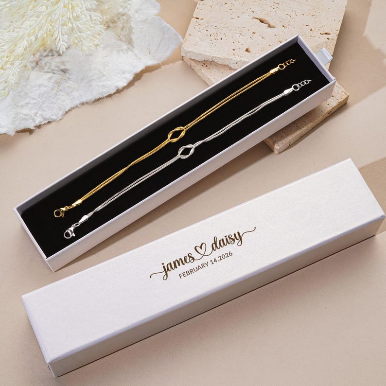 K&ouml;nnte beinhalten: Zwei feine Armb&auml;nder, eines goldfarben und eines silberfarben, werden in einer schwarz gef&uuml;tterten wei&szlig;en Schachtel pr&auml;sentiert. Jedes Armband hat ein Knotendesign in der Mitte. Die Schachtel ist ge&ouml;ffnet und gibt den Schmuck frei. Der Deckel tr&auml;gt den Text "james ♡ daisy FEBRUARY 14.2026".