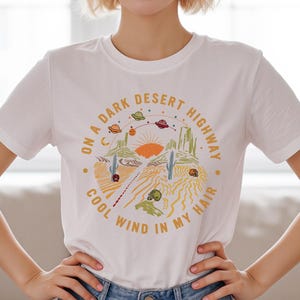 May include: White t-shirt featuring a desert landscape design with the text "ON A DARK DESERT HIGHWAY COOL WIND IN MY HAIR." The graphic includes a sun, cacti, and UFOs. The shirt is a short-sleeved crew neck.