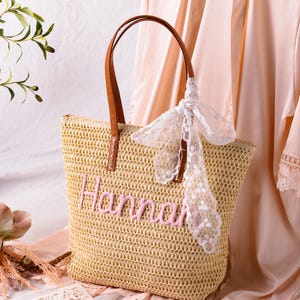 May include: A woven straw tote bag featuring the name Hannah embroidered in pink thread on the front. The bag has two brown leather-like handles and is decorated with a white lace ribbon tied to one handle. The texture is a natural tan colour.