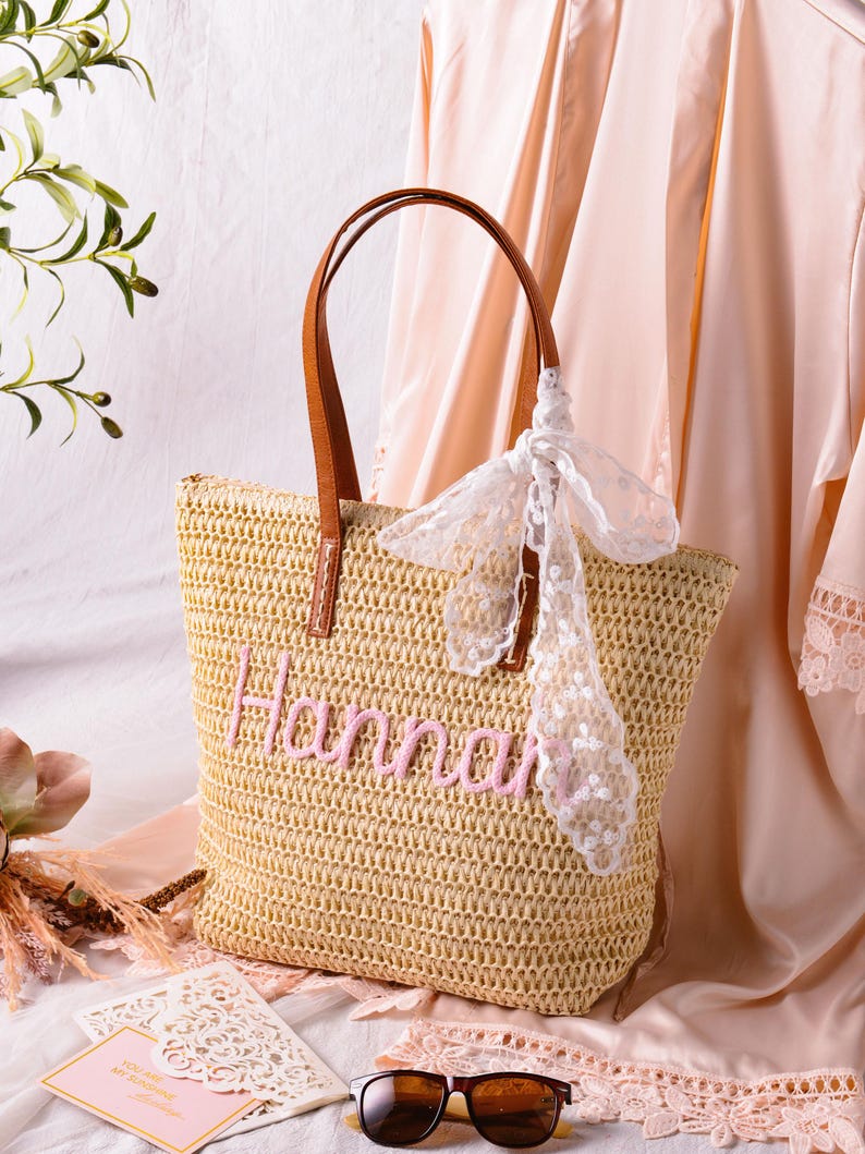 May include: A woven straw tote bag featuring the name Hannah embroidered in pink thread on the front. The bag has two brown leather-like handles and is decorated with a white lace ribbon tied to one handle. The texture is a natural tan colour.