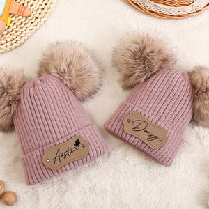 Puede incluir: Dos gorros de punto rosa con grandes pompones de piel sintética. Cada gorro tiene un parche de polipiel marrón con un nombre. Uno dice "Austin" y el otro "Daisy".