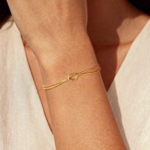 K&ouml;nnte beinhalten: Ein filigranes goldenes Armband mit einem Knotendesign. Das Armband ist d&uuml;nn und elegant, mit einem schlichten, aber stilvollen Aussehen. Die goldene Farbe verleiht dem Schmuckst&uuml;ck einen Hauch von Luxus, passend f&uuml;r den Alltag oder besondere Anl&auml;sse.
