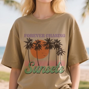 May include: A tan t-shirt with a graphic of a sunset scene. The design includes palm trees, an orange sun, and the words "FOREVER CHASING SUNSETS" in purple and green. The shirt has short sleeves.