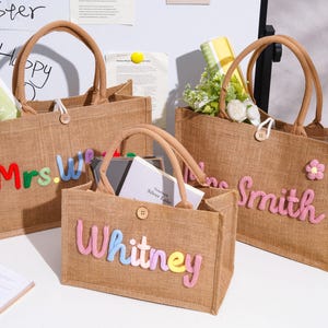May include: Three rectangular jute tote bags with brown handles and a single wooden button closure. The bags feature colorful, raised, embroidered lettering on the front. One bag displays "Mrs.W", another "Whitney", and the third "Smith" with a flower.
