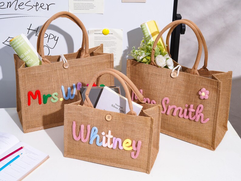 May include: Three rectangular jute tote bags with brown handles and a single wooden button closure. The bags feature colorful, raised, embroidered lettering on the front. One bag displays "Mrs.W", another "Whitney", and the third "Smith" with a flower.