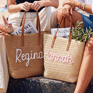 May include: Two woven straw tote bags with brown leather handles. The left bag is tan with the name Regina embroidered in white script. The right bag is light beige with the name Hannah embroidered in pink script. Both bags contain magazines and greenery.