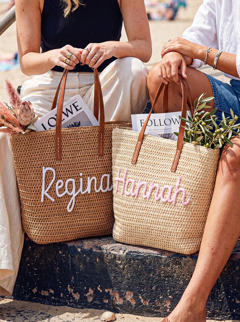 May include: Two woven straw tote bags with brown leather handles. The left bag is tan with the name Regina embroidered in white script. The right bag is light beige with the name Hannah embroidered in pink script. Both bags contain magazines and greenery.