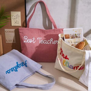 K&ouml;nnte beinhalten: Drei Tragetaschen: eine rosa Tasche mit wei&szlig;em Sticktext Best Teacher, eine hellblaue Tasche mit blauem Sticktext Angelina und eine cremefarbene Tasche mit mehrfarbigem Sticktext Mrs. Williams.