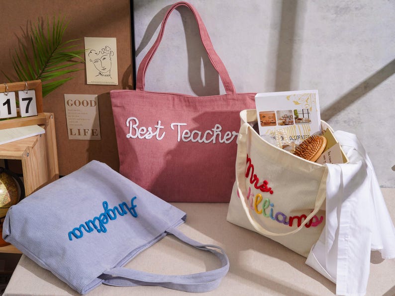 K&ouml;nnte beinhalten: Drei Tragetaschen: eine rosa Tasche mit wei&szlig;em Sticktext Best Teacher, eine hellblaue Tasche mit blauem Sticktext Angelina und eine cremefarbene Tasche mit mehrfarbigem Sticktext Mrs. Williams.