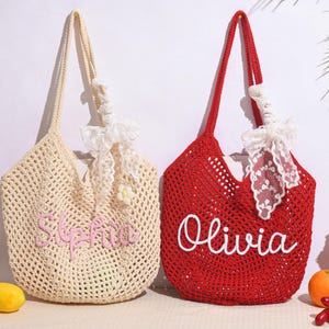 May include: Two crochet tote bags. One is cream-colored with the name "Sophia" embroidered in pink. The other is red with the name "Olivia" in white. Both bags have long handles and are decorated with white lace and bows.