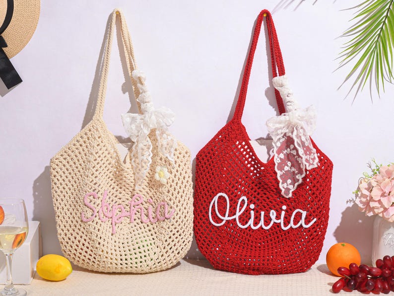 May include: Two crochet tote bags. One is cream-colored with the name "Sophia" embroidered in pink. The other is red with the name "Olivia" in white. Both bags have long handles and are decorated with white lace and bows.