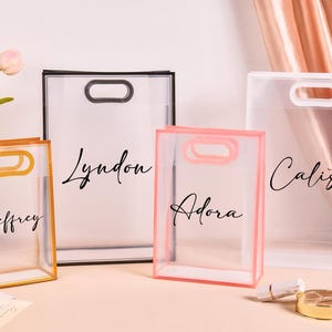 Könnte beinhalten: Vier transparente Tragetaschen mit unterschiedlich farbigen Rahmen: Gold, Schwarz und Rosa. Jede Tasche hat einen Griff und einen aufgedruckten Namen: Jeffrey, Lyndon, Adora und Calista. Ein Lippenstift und ein Kompaktpuder befinden sich im Vordergrund.