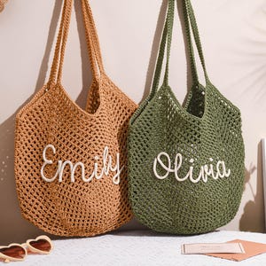 May include: Two open-weave crochet tote bags. One is tan with the name "Emily" in white script, the other is olive green with "Olivia". Both have long handles and are hanging against a neutral wall.