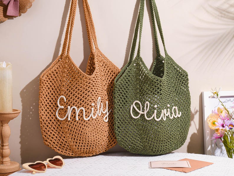 May include: Two open-weave crochet tote bags. One is tan with the name "Emily" in white script, the other is olive green with "Olivia". Both have long handles and are hanging against a neutral wall.