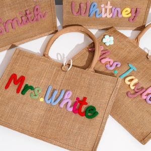 May include: A collection of rectangular burlap tote bags with two handles and a wooden button closure. Each bag features personalized names in colorful, raised, yarn-wrapped lettering, including Mrs. Smith, Whitney, Mrs. White, and Mrs. Taylor.