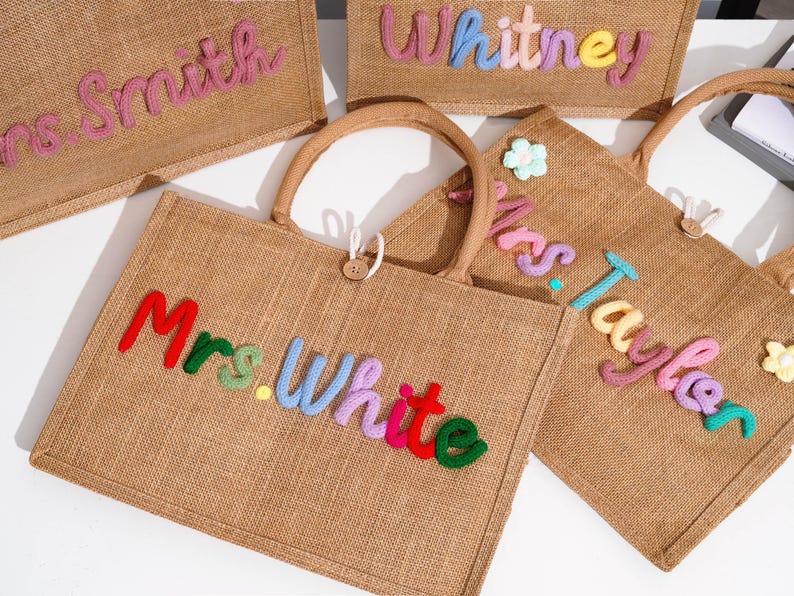 May include: A collection of rectangular burlap tote bags with two handles and a wooden button closure. Each bag features personalized names in colorful, raised, yarn-wrapped lettering, including Mrs. Smith, Whitney, Mrs. White, and Mrs. Taylor.