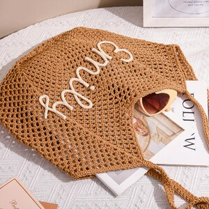 May include: A brown, open-weave tote bag with the name "Emily" in white script. The bag contains a pair of heart-shaped sunglasses and a book. The bag has long, braided handles and is sitting on a white surface.