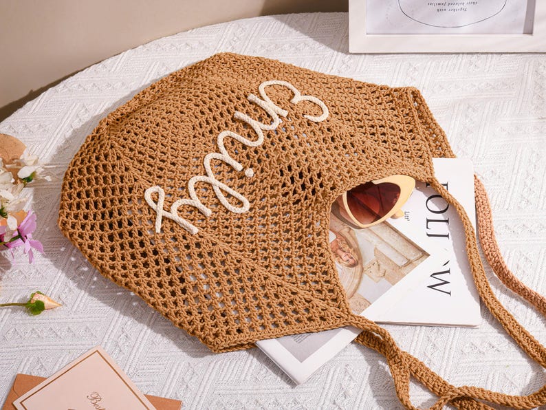 May include: A brown, open-weave tote bag with the name "Emily" in white script. The bag contains a pair of heart-shaped sunglasses and a book. The bag has long, braided handles and is sitting on a white surface.