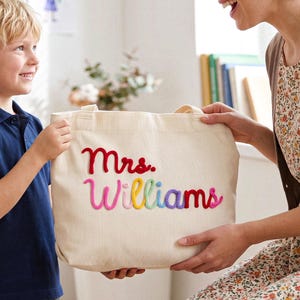 Può includere: Borsa tote color crema a righe verticali. La borsa presenta il nome "Mrs. Williams" ricamato con filo colorato. La borsa è tenuta da una donna e da un bambino.