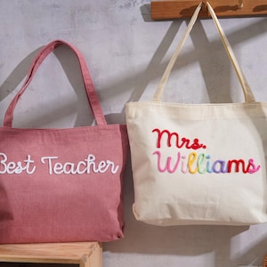 K&ouml;nnte beinhalten: Zwei Cord-Einkaufstaschen mit Schulterriemen. Die linke Tasche ist altrosa mit wei&szlig;em gesticktem Text Best Teacher. Die rechte Tasche ist cremefarben mit mehrfarbigem gesticktem Text Mrs. Williams in Rot, Pink, Gelb und Blau.