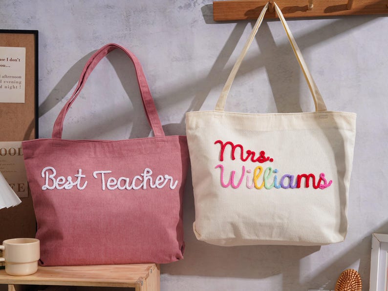 K&ouml;nnte beinhalten: Zwei Cord-Einkaufstaschen mit Schulterriemen. Die linke Tasche ist altrosa mit wei&szlig;em gesticktem Text Best Teacher. Die rechte Tasche ist cremefarben mit mehrfarbigem gesticktem Text Mrs. Williams in Rot, Pink, Gelb und Blau.