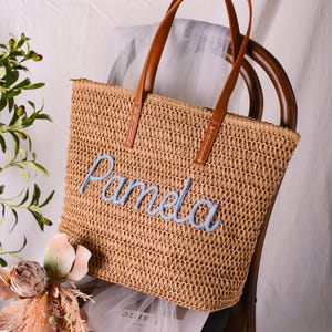 May include: A tan woven straw tote bag featuring two brown leather handles and the name Pamela embroidered in light blue script across the front. The bag has a rectangular shape with a textured, open-weave construction and a top zipper closure.
