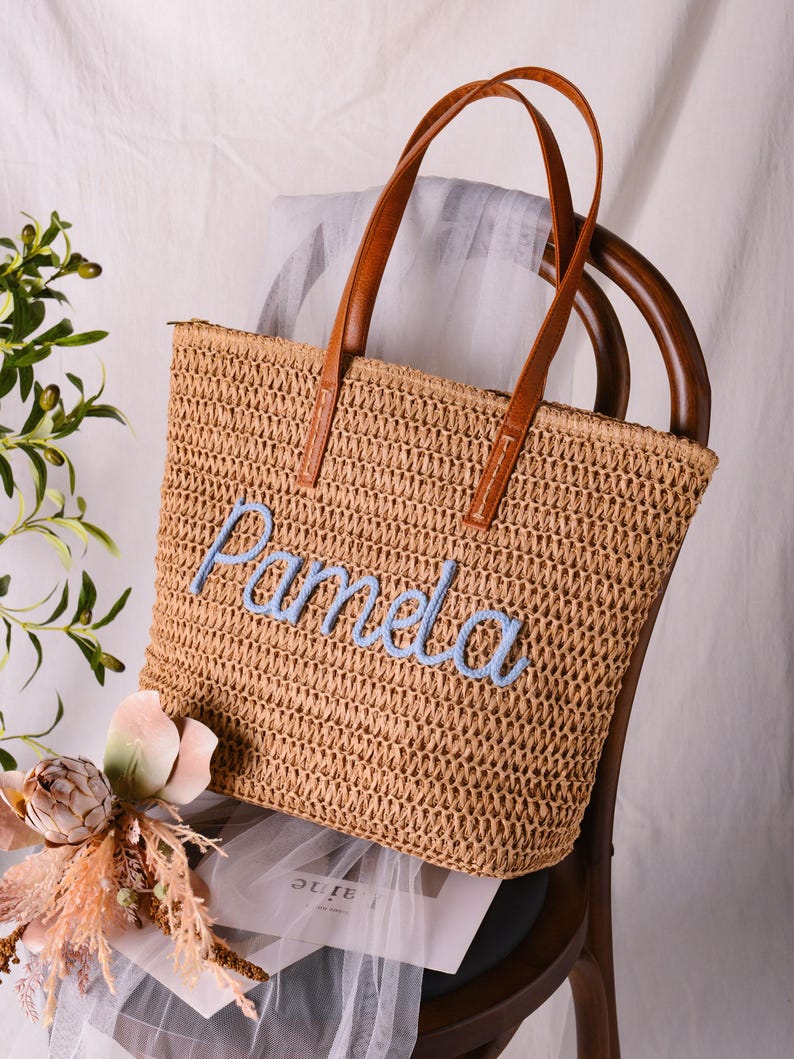 May include: A tan woven straw tote bag featuring two brown leather handles and the name Pamela embroidered in light blue script across the front. The bag has a rectangular shape with a textured, open-weave construction and a top zipper closure.