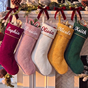 Puede incluir: Cinco calcetines navideños personalizados de tela acanalada en rojo oscuro, rosa claro, blanco, amarillo mostaza y verde oscuro. Cada calcetín tiene un puño con un nombre bordado en letra blanca: Sophia, Kristin, Olivia, Nicole y Keith.