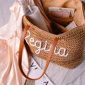 May include: A woven straw tote bag with brown leather handles and the name Regina embroidered in white script on the front. The open bag contains a white tumbler with a straw, a small card, and a tube, while a pale pink satin fabric rests nearby.