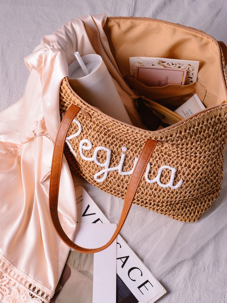 May include: A woven straw tote bag with brown leather handles and the name Regina embroidered in white script on the front. The open bag contains a white tumbler with a straw, a small card, and a tube, while a pale pink satin fabric rests nearby.