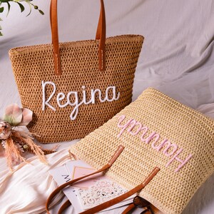 May include: Two woven straw tote bags with brown leather-look handles. One bag is tan with the name Regina embroidered in white script, and the other is a lighter straw colour with the name Hannah embroidered in pink script on the front surface.