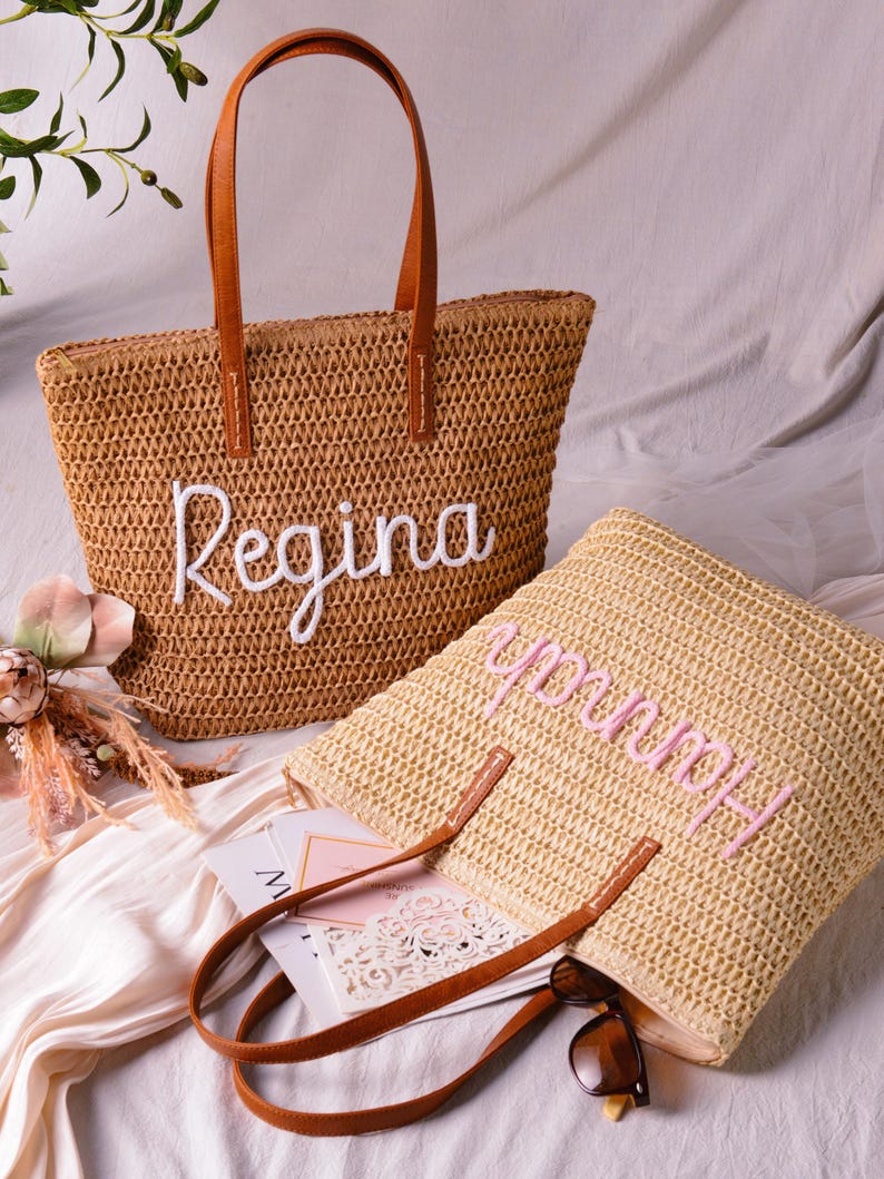 May include: Two woven straw tote bags with brown leather-look handles. One bag is tan with the name Regina embroidered in white script, and the other is a lighter straw colour with the name Hannah embroidered in pink script on the front surface.