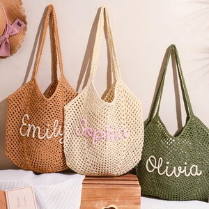 May include: Three crochet tote bags in shades of brown, cream, and green, each with a name embroidered on the front. The bags have long handles and a woven design. A straw hat with a pink bow is visible in the background.