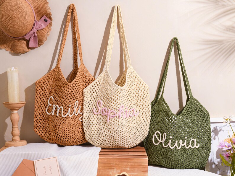 May include: Three crochet tote bags in shades of brown, cream, and green, each with a name embroidered on the front. The bags have long handles and a woven design. A straw hat with a pink bow is visible in the background.