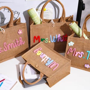 May include: Four rectangular brown jute tote bags with two handles and a single button closure. Each bag features colorful, raised, fabric-stitched names including Mrs. Smith, Mrs. White, Mrs. Taylor, and Whitney, accented with small fabric flower appliques.