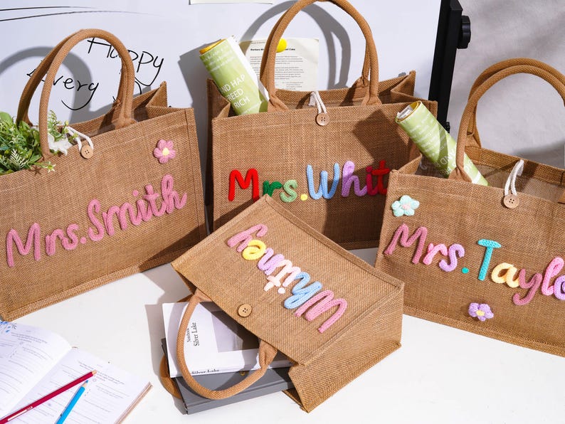 May include: Four rectangular brown jute tote bags with two handles and a single button closure. Each bag features colorful, raised, fabric-stitched names including Mrs. Smith, Mrs. White, Mrs. Taylor, and Whitney, accented with small fabric flower appliques.