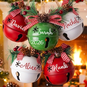 May include: A set of five round metal jingle bell ornaments in red, green, and white. Each bell features a name in script font, including David, Rachel, Sarah, Alice, and Hunk, with the year 2026 on the green bell. Each is topped with pine, berries, and a red gingham bow.